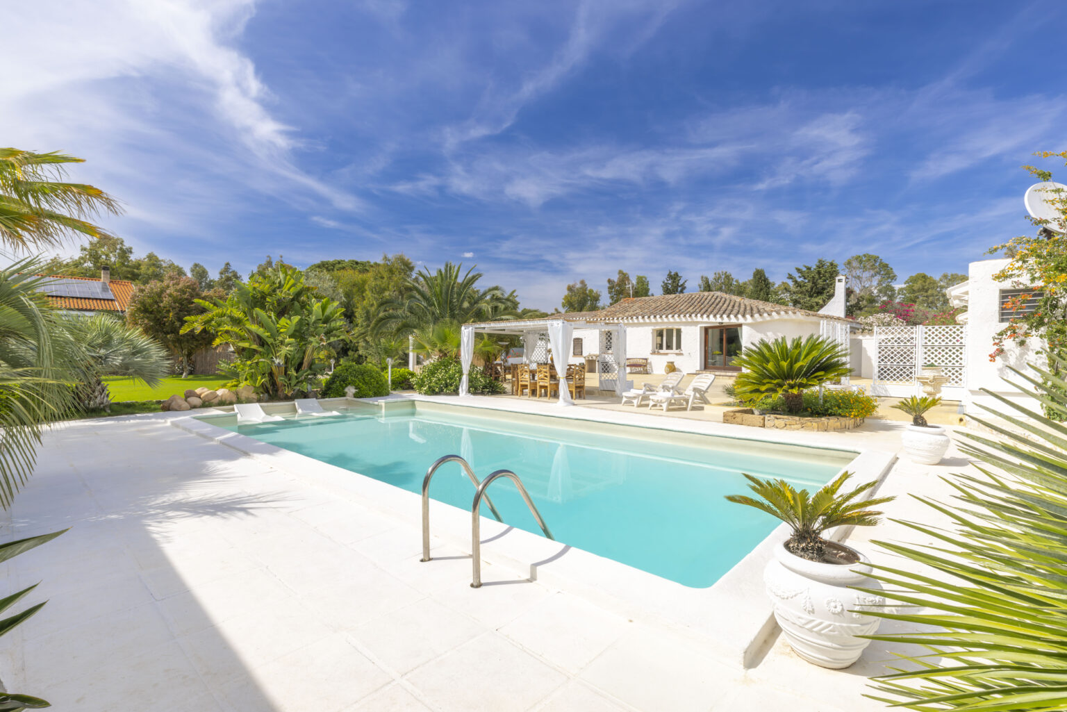 Luxurious outdoor swimming pool surrounded by lush greenery and a modern villa, featuring sun loungers and a clear blue sky. Ideal for relaxation and leisure activities. | Only on SopranoVillas