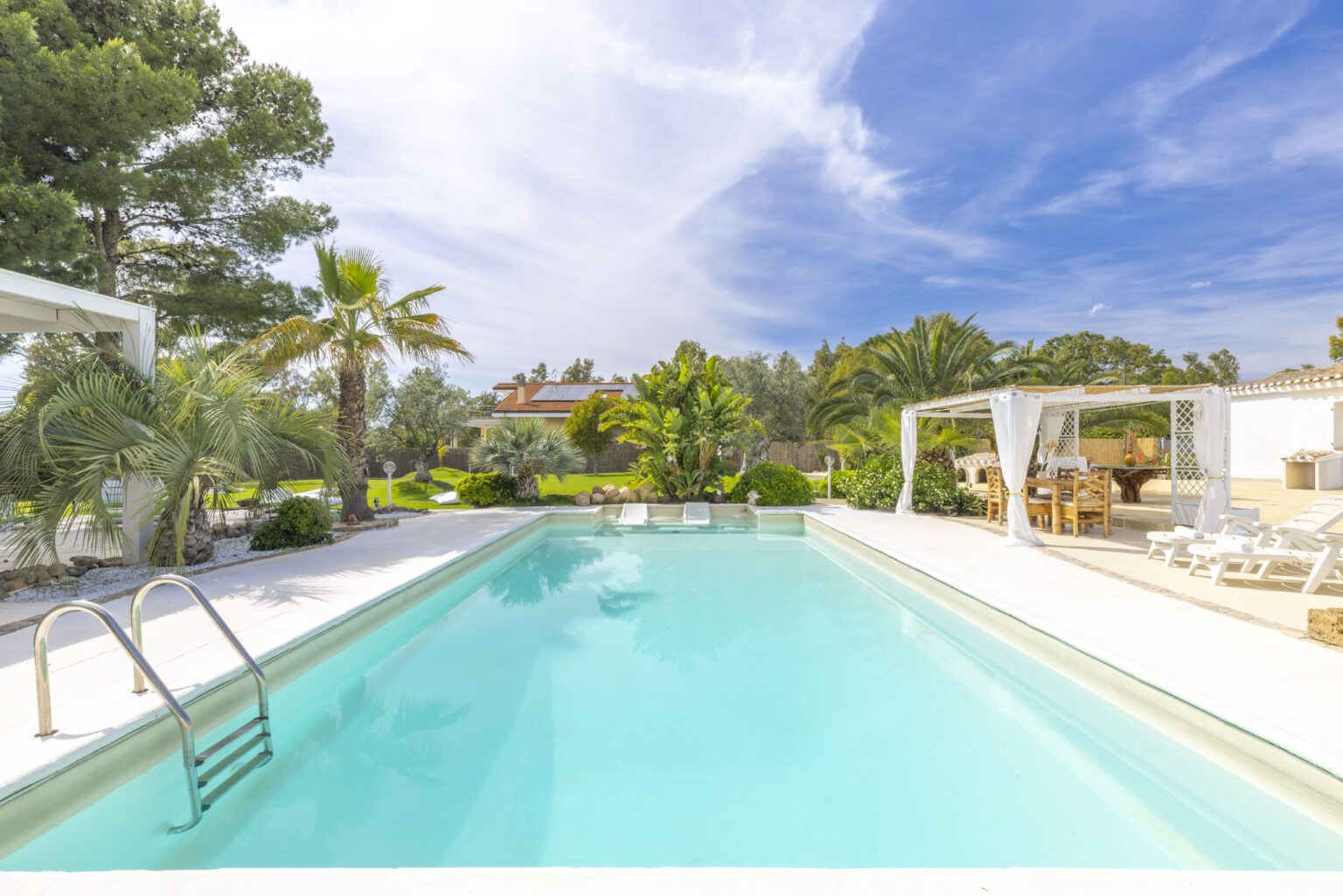 Luxury villa with a pristine swimming pool surrounded by palm trees and lush greenery, featuring a shaded outdoor dining area and sun loungers, ideal for relaxation and entertaining in a tropical setting. | Only on SopranoVillas