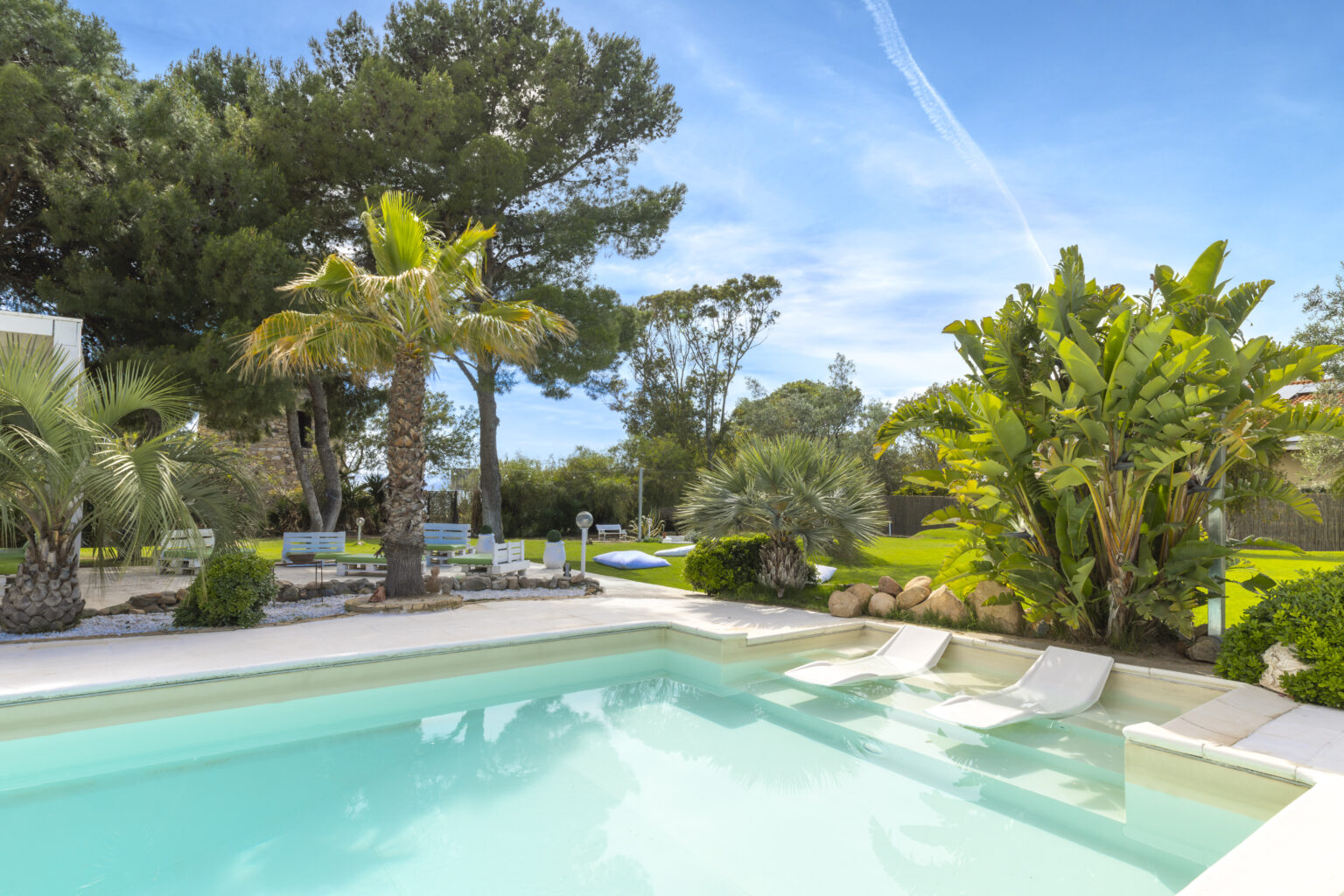Luxurious outdoor swimming pool surrounded by lush greenery, palm trees, and tropical plants under a clear blue sky, ideal for relaxation and leisure. | Only on SopranoVillas