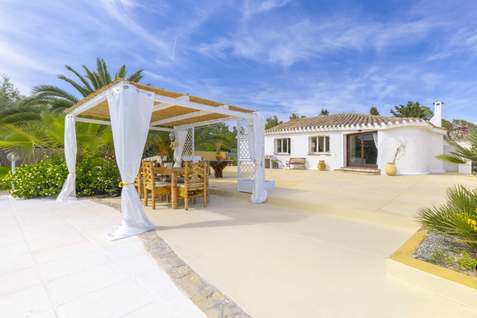 Charming outdoor patio with a bamboo dining set under a pergola, surrounded by lush greenery and a white-walled house, set against a bright blue sky. Ideal for relaxation and outdoor dining. | Only on SopranoVillas