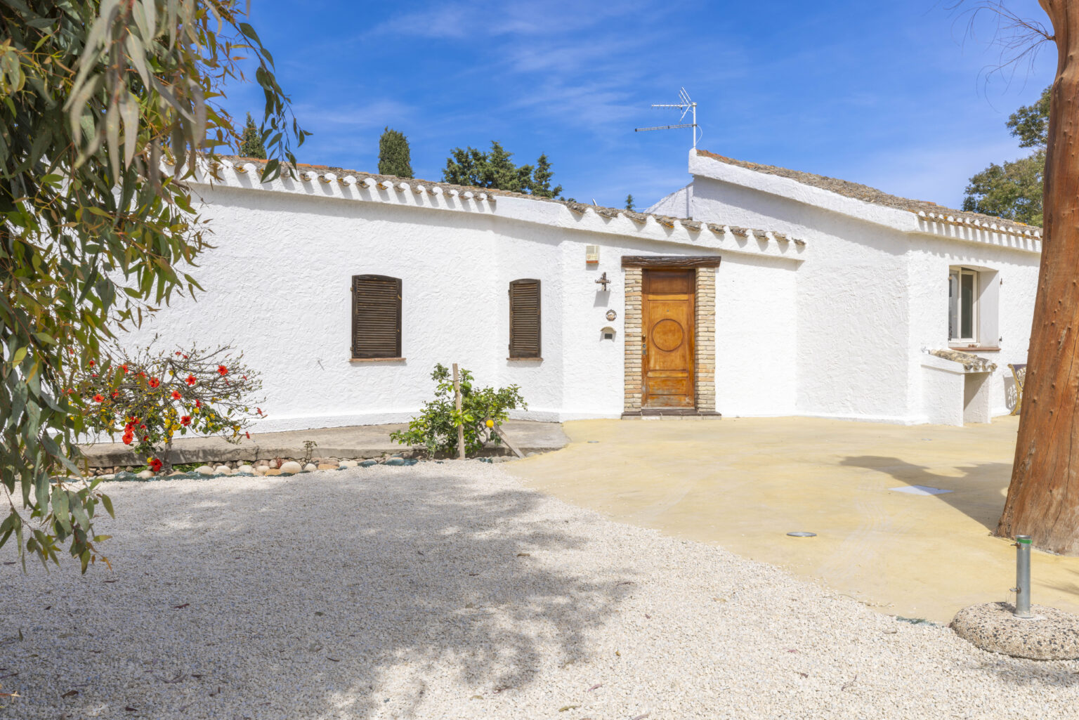Charming white house with a wooden door, surrounded by landscaped gardens and gravel driveway, under a clear blue sky. Ideal for showcasing residential properties or vacation homes. | Only on SopranoVillas