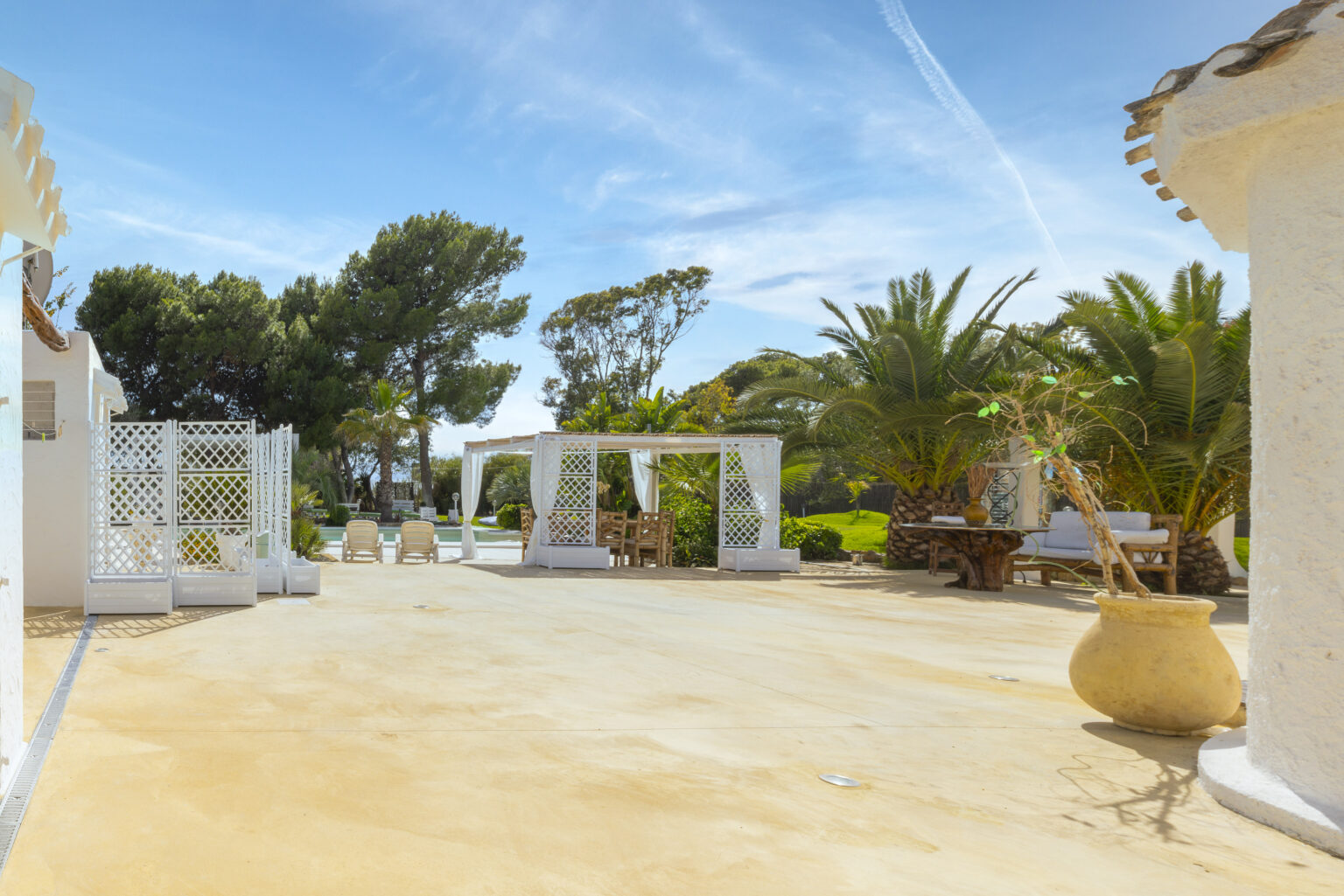 Tropical outdoor patio area featuring white architectural structures, lush palm trees, and a serene blue sky, ideal for relaxation and gatherings. | Only on SopranoVillas