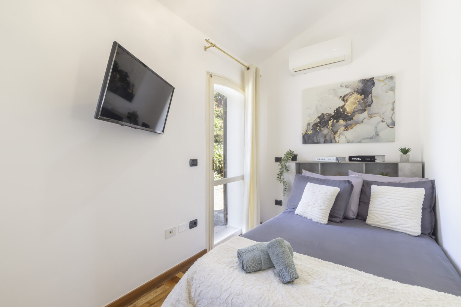 Cozy modern bedroom featuring a queen-sized bed with gray bedding, a wall-mounted TV, and a large window for natural light. Decor includes a contemporary artwork piece and indoor plants, creating a relaxing atmosphere. | Only on SopranoVillas