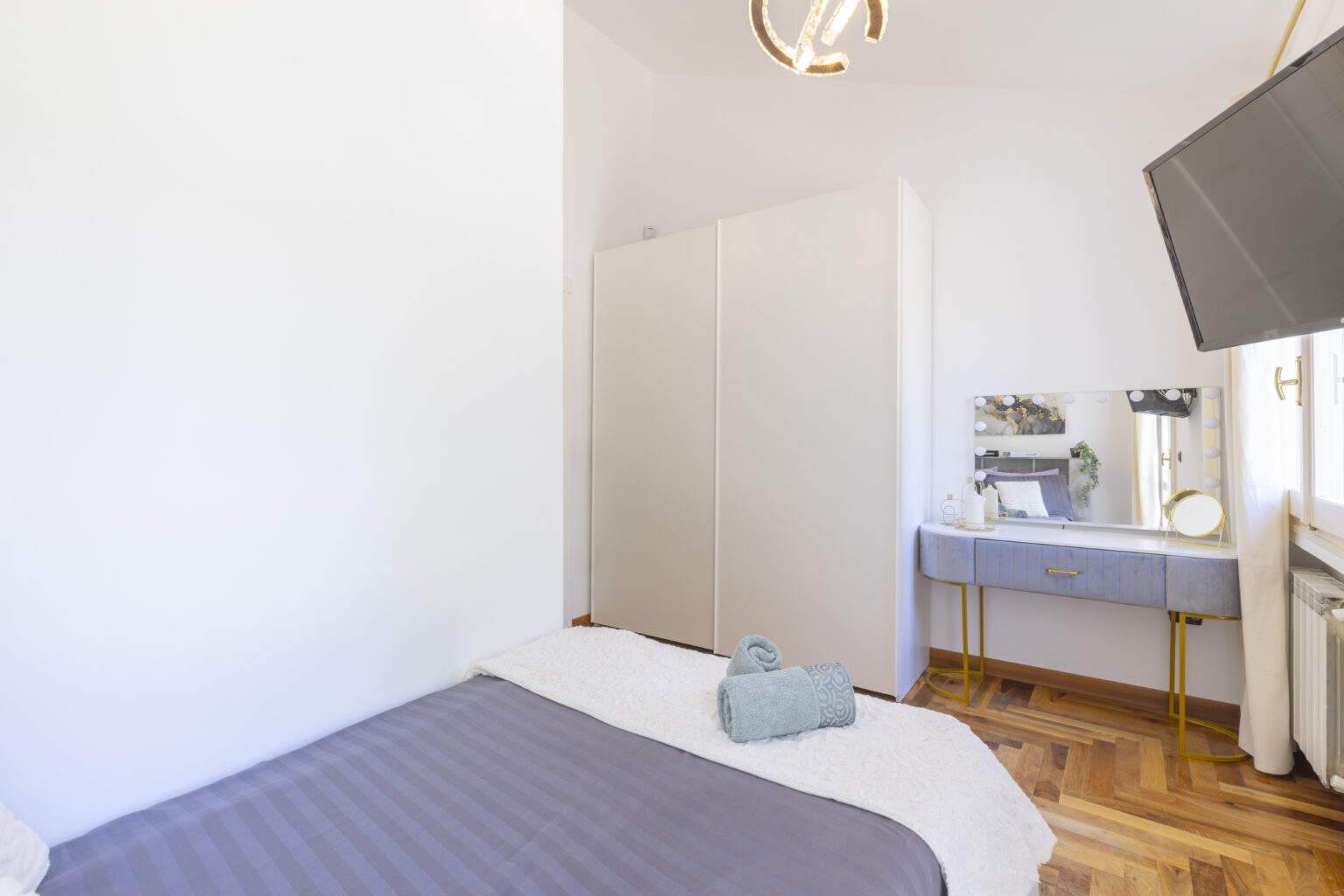 Cozy bedroom featuring a gray bedspread, wooden flooring, a modern vanity with a mirror, and a wall-mounted television, illuminated by natural light from a nearby window. | Only on SopranoVillas