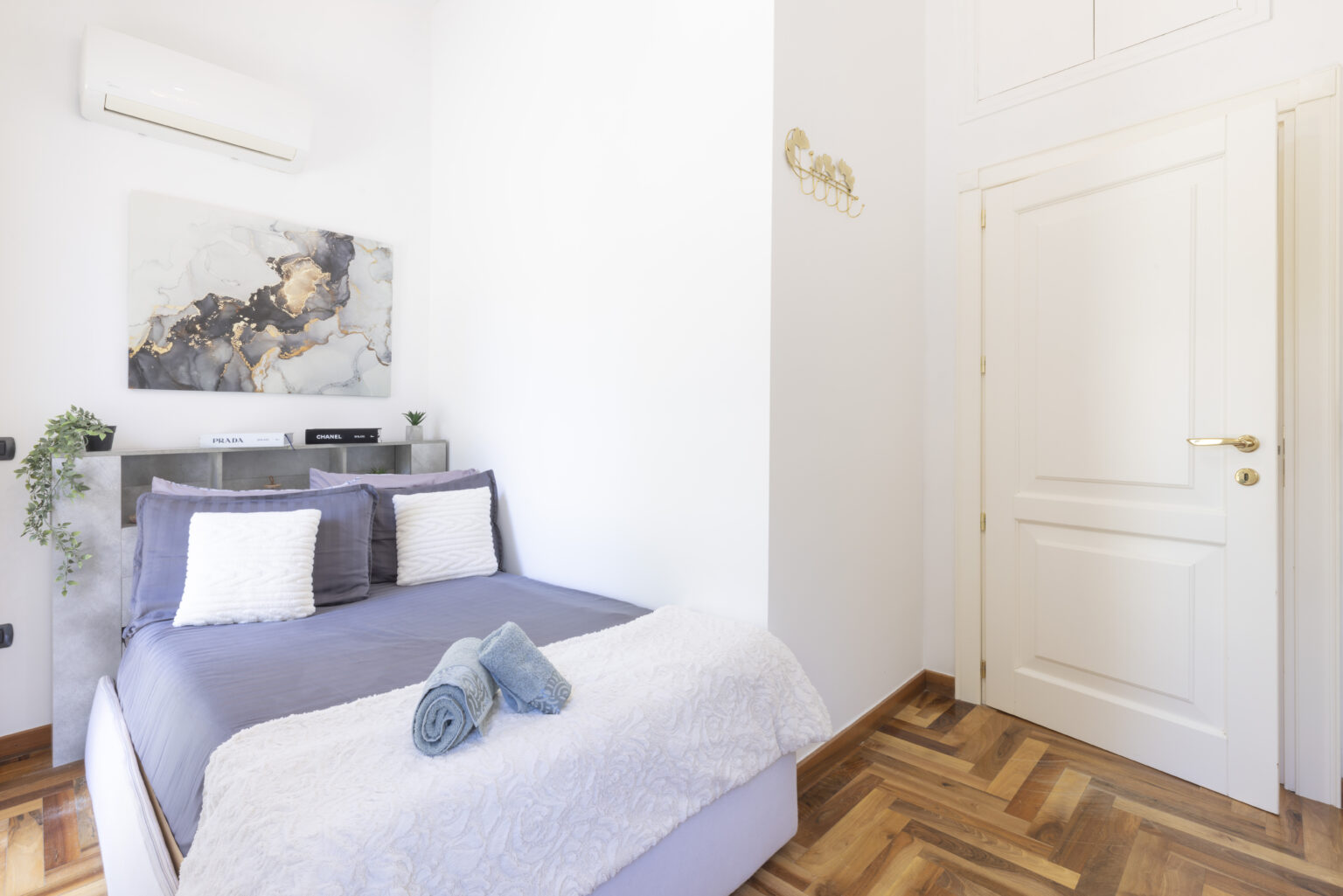Cozy bedroom with a double bed, featuring a modern aesthetic, light-colored walls, and wooden flooring. The room includes decorative pillows, a soft blanket, and a piece of artwork on the wall, complemented by an air conditioning unit and a stylish door. Ideal for comfortable living or guest accommodation. | Only on SopranoVillas