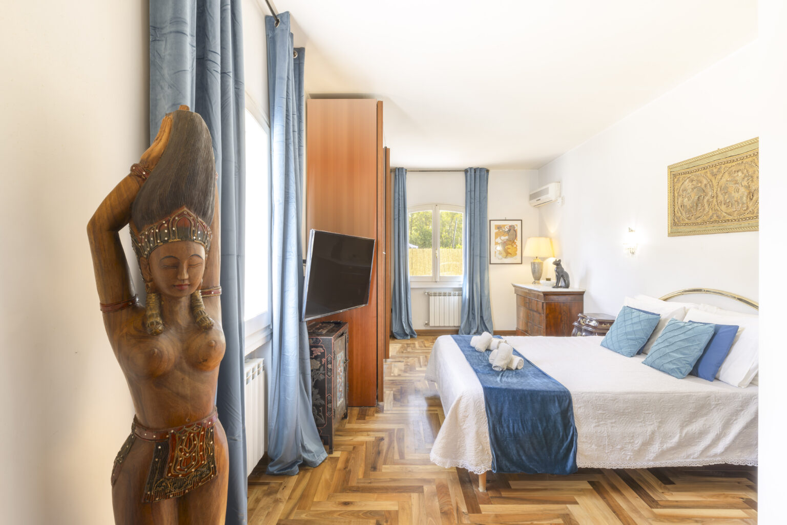 Cozy and stylish bedroom featuring wooden flooring, elegant blue curtains, a decorative wooden statue, and a comfortable bed with blue and white linens. The room includes a television and a wooden dresser, creating a warm and inviting atmosphere. Ideal for relaxation and comfort. | Only on SopranoVillas