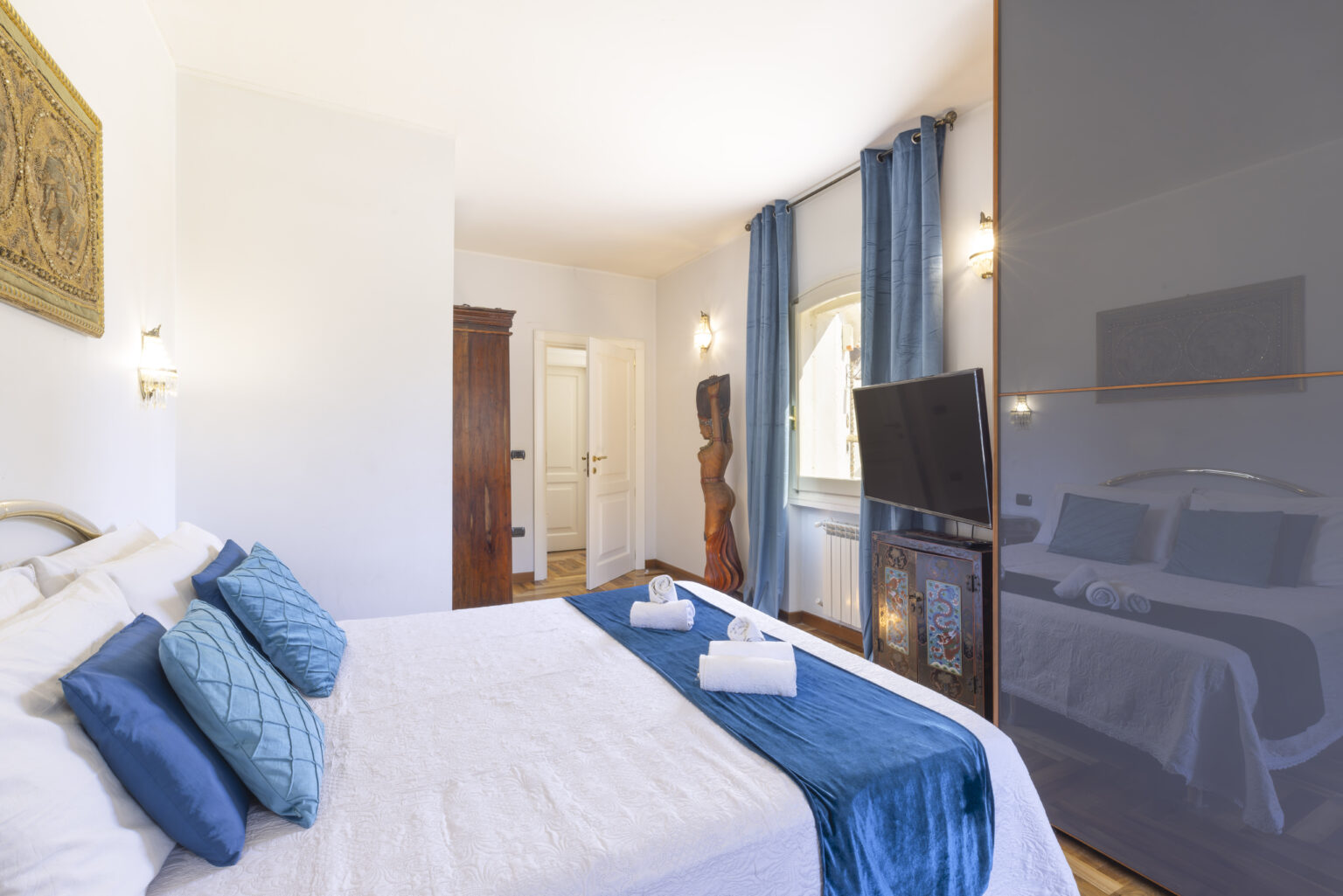 Modern bedroom with a comfortable queen-sized bed adorned with blue decorative pillows, featuring a stylish TV unit and large windows with blue curtains, creating a bright and inviting atmosphere. | Only on SopranoVillas