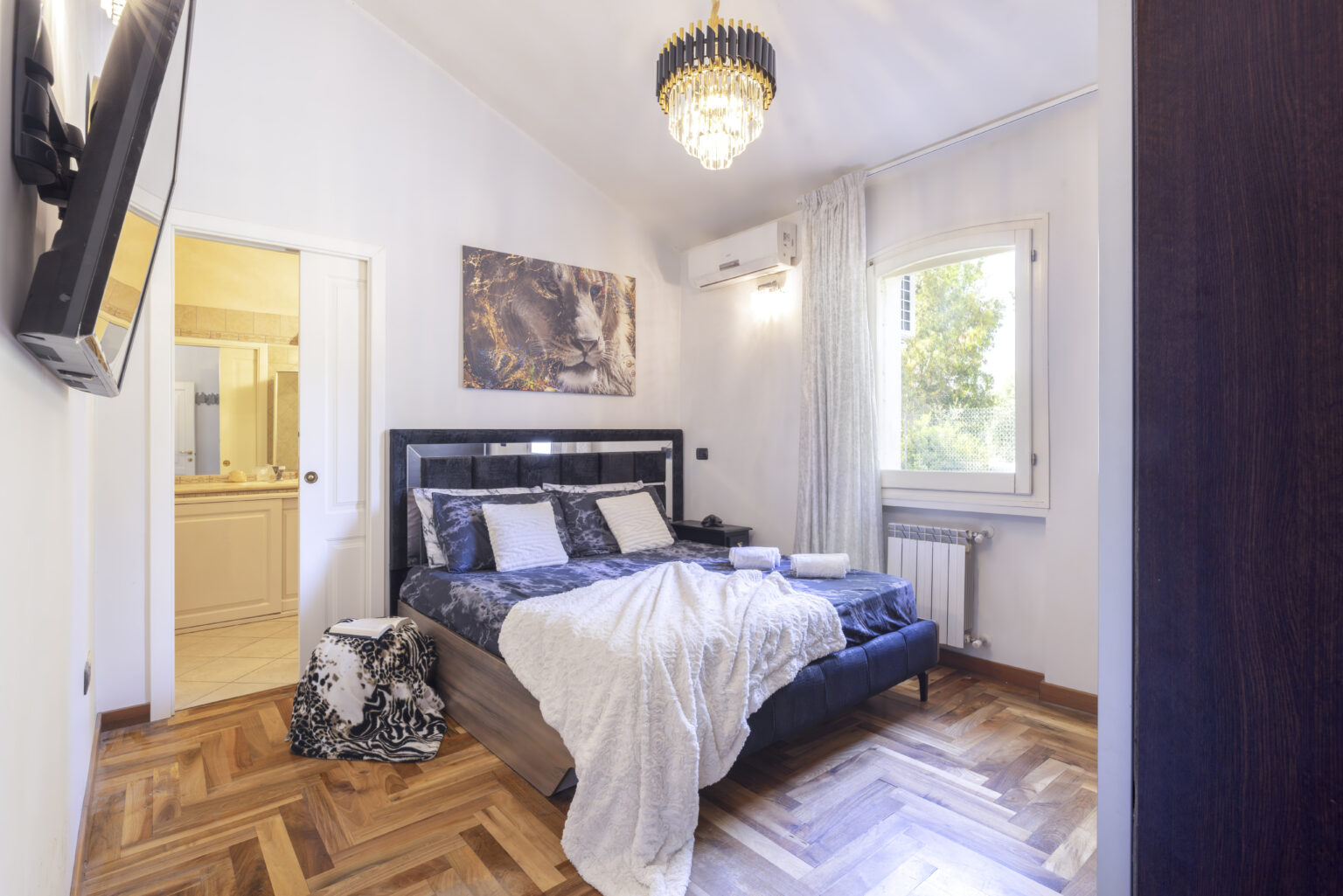 Modern bedroom with a stylish black bed, decorative pillows, and a cozy white throw blanket. Features a chandelier, a wall-mounted TV, and a view of greenery through large windows. Adjacent bathroom visible through an open door, showcasing elegant decor and tile work. Warm wooden flooring enhances the inviting atmosphere. | Only on SopranoVillas