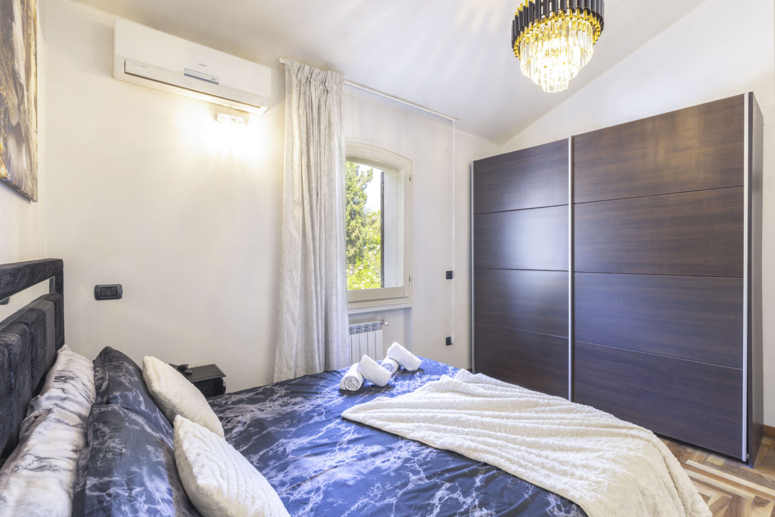 Modern bedroom featuring a comfortable bed with plush bedding, a stylish wardrobe, and natural light from a window, complemented by elegant decor and a chandelier. Ideal for relaxation and comfort. | Only on SopranoVillas