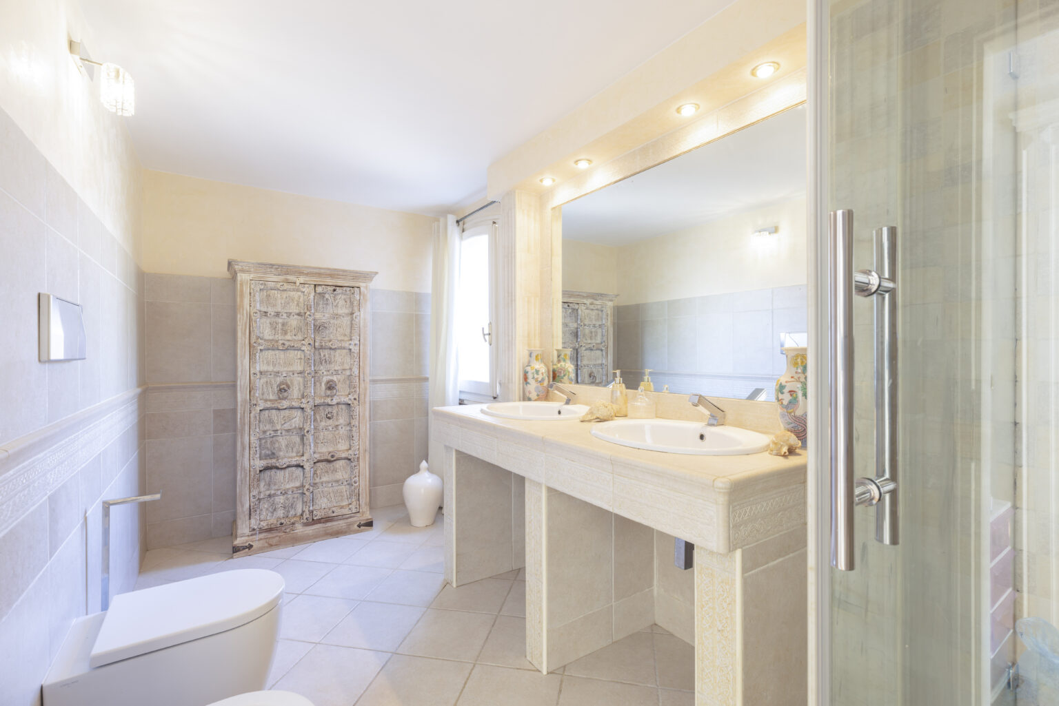 Modern bathroom featuring a double sink vanity, elegant fixtures, a decorative wooden cabinet, and a glass shower enclosure. Brightly lit with natural light, the neutral color palette enhances the spacious feel. | Only on SopranoVillas