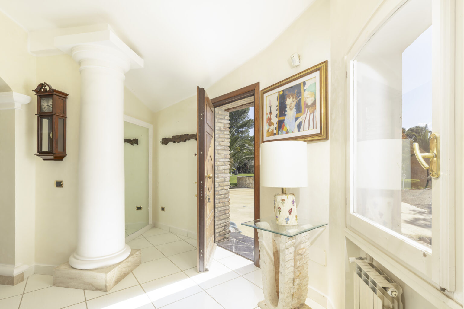 Bright and inviting entryway featuring a decorative column, a wooden door leading to the outdoors, a stylish lamp on a marble table, and artwork on the wall, creating a welcoming atmosphere. | Only on SopranoVillas