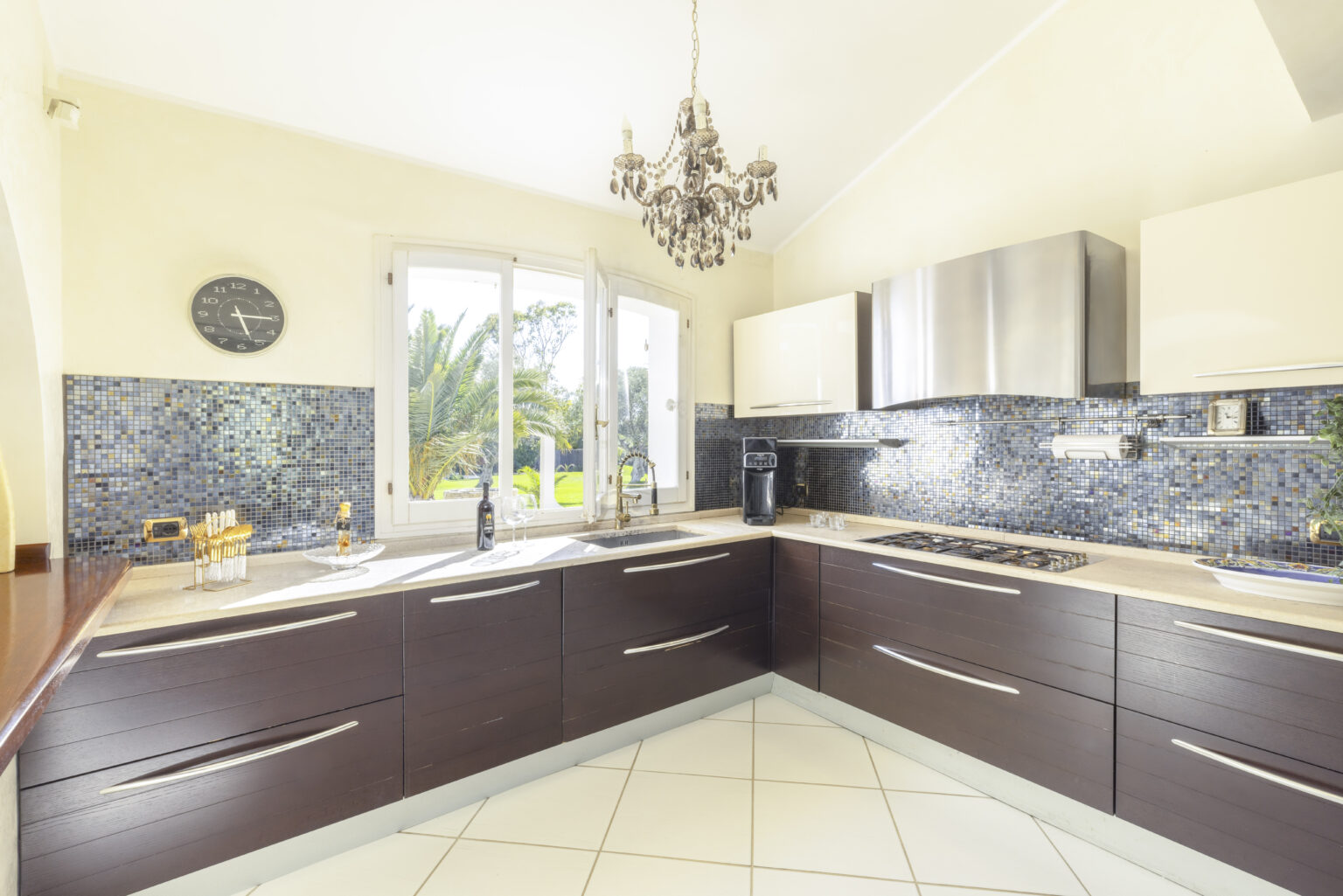 Modern kitchen featuring dark wooden cabinets, stainless steel appliances, and a decorative chandelier. Bright natural light streams in through large windows, highlighting the stylish tile flooring and blue mosaic backsplash. | Only on SopranoVillas