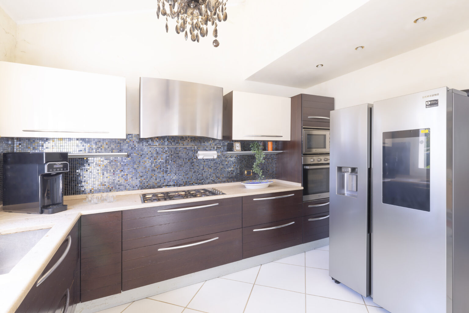 Modern kitchen with dark wood cabinetry, stainless steel appliances, and a stylish chandelier. Features a sleek countertop, built-in oven, and contemporary tile flooring, creating an elegant cooking space. | Only on SopranoVillas