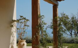A serene view from a rustic porch featuring wooden beams and potted plants, with a backdrop of blue sky and distant greenery by the sea.