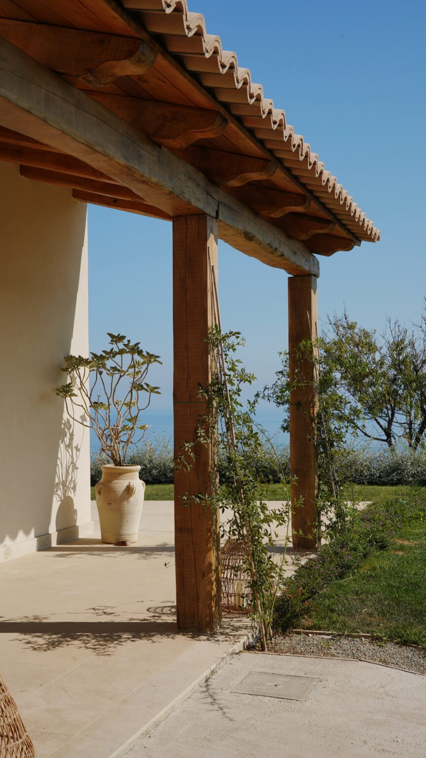 A serene view from a rustic porch featuring wooden beams and potted plants, with a backdrop of blue sky and distant greenery by the sea. | Only on SopranoVillas