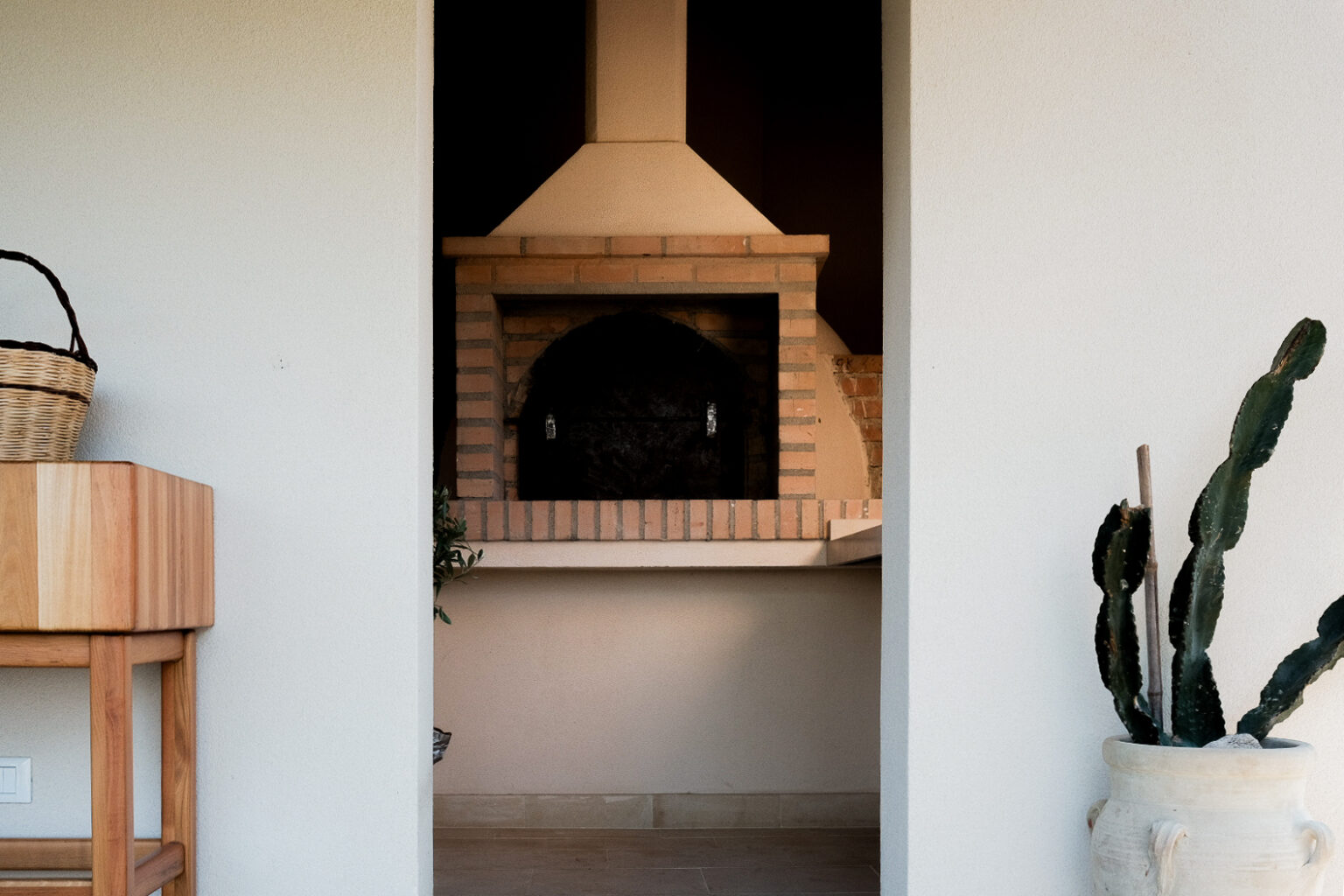 Cozy outdoor space featuring a rustic brick pizza oven, with a wooden table and potted cactus in the foreground. Ideal for alfresco dining and entertaining. | Only on SopranoVillas