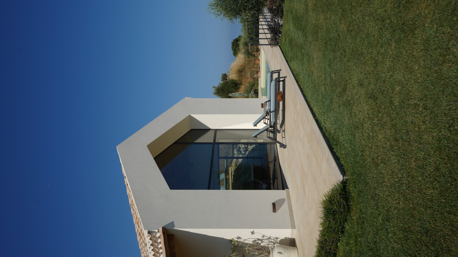 Modern architectural design of a house with large glass windows, featuring a patio area with lounge chairs, a well-maintained lawn, and a clear blue sky in the background. Ideal for outdoor relaxation and enjoying scenic views. | Only on SopranoVillas