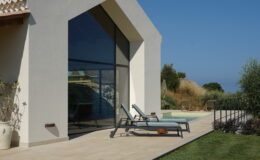 Modern architectural design of a house with large glass windows, featuring a patio area with lounge chairs, a well-maintained lawn, and a clear blue sky in the background. Ideal for outdoor relaxation and enjoying scenic views.