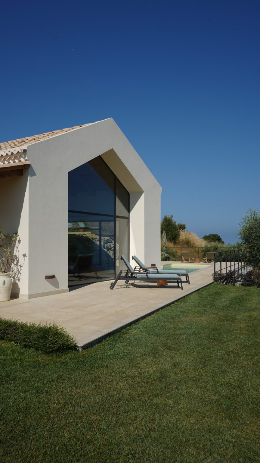 Modern architectural design of a house with large glass windows, featuring a patio area with lounge chairs, a well-maintained lawn, and a clear blue sky in the background. Ideal for outdoor relaxation and enjoying scenic views. | Only on SopranoVillas