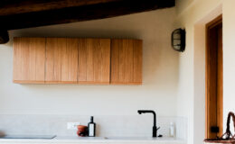 Modern kitchen interior featuring wooden upper cabinets, a sleek black faucet, and a minimalist countertop with a bottle and decorative items. Natural light highlights the warm tones of the wood and the clean lines of the design.