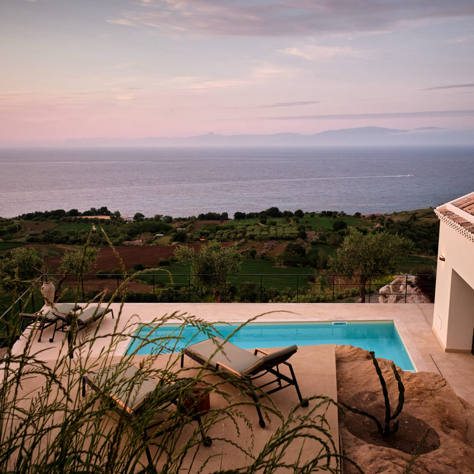 Scenic view of a modern villa with a swimming pool overlooking the ocean and lush green landscape at sunset. | Only on SopranoVillas
