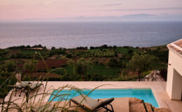Scenic view of a modern villa with a swimming pool overlooking the ocean and lush green landscape at sunset.