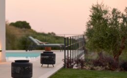 Outdoor patio with modern fire pits, a swimming pool, and a lounge chair against a serene sunset backdrop, surrounded by lush greenery and a stylish fence.
