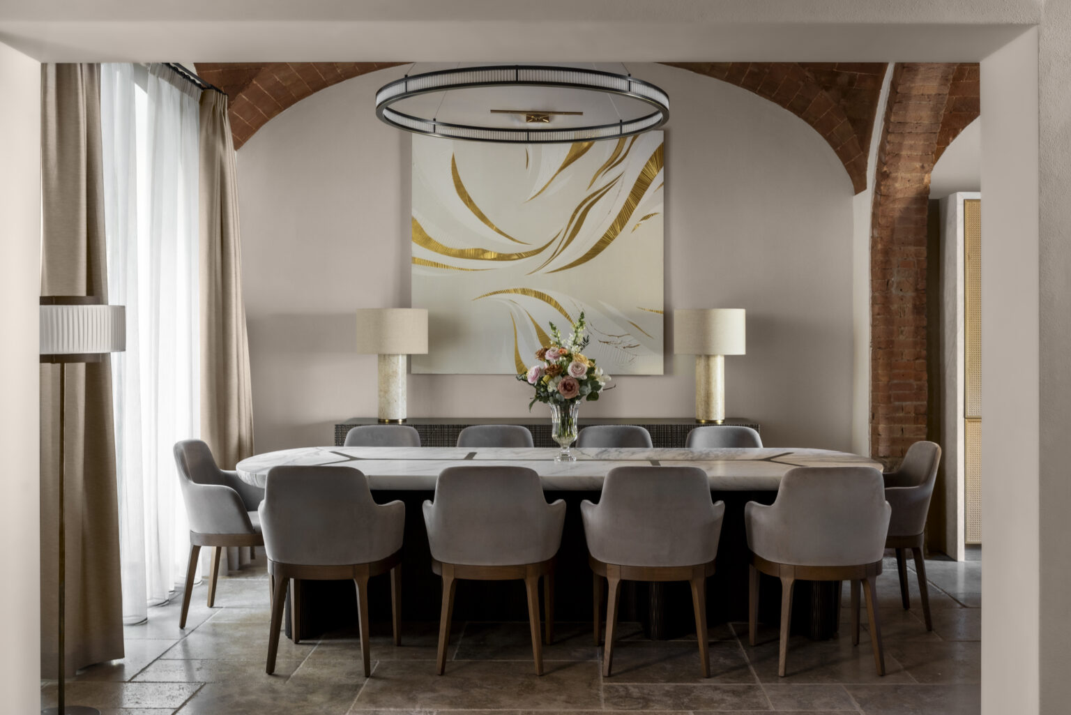 Modern dining room featuring a large oval table surrounded by gray upholstered chairs, complemented by stylish lamps and a vibrant abstract painting on the wall, illuminated by a contemporary circular chandelier. | Only on SopranoVillas