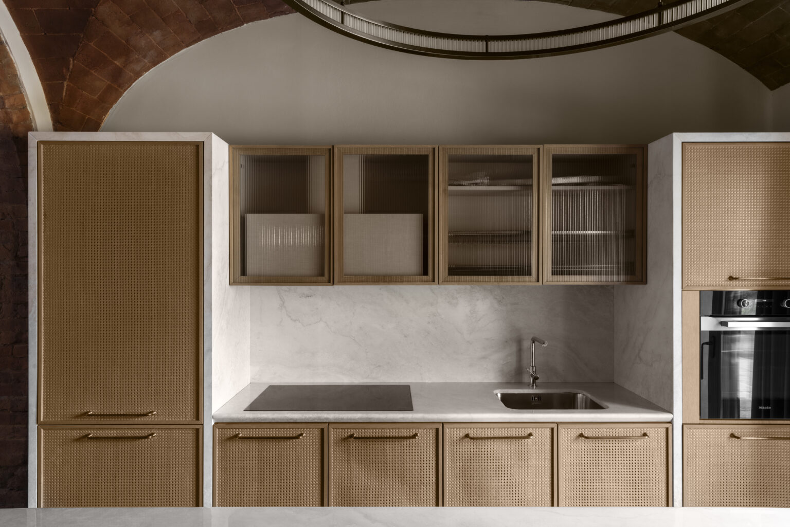 Modern kitchen design featuring a sleek layout with a minimalist aesthetic. The cabinetry combines natural wood finishes with a textured pattern, complemented by a marble countertop. A stainless steel sink and cooktop are integrated into the space, enhancing functionality and style. The arching ceiling adds architectural interest, creating an inviting atmosphere. | Only on SopranoVillas