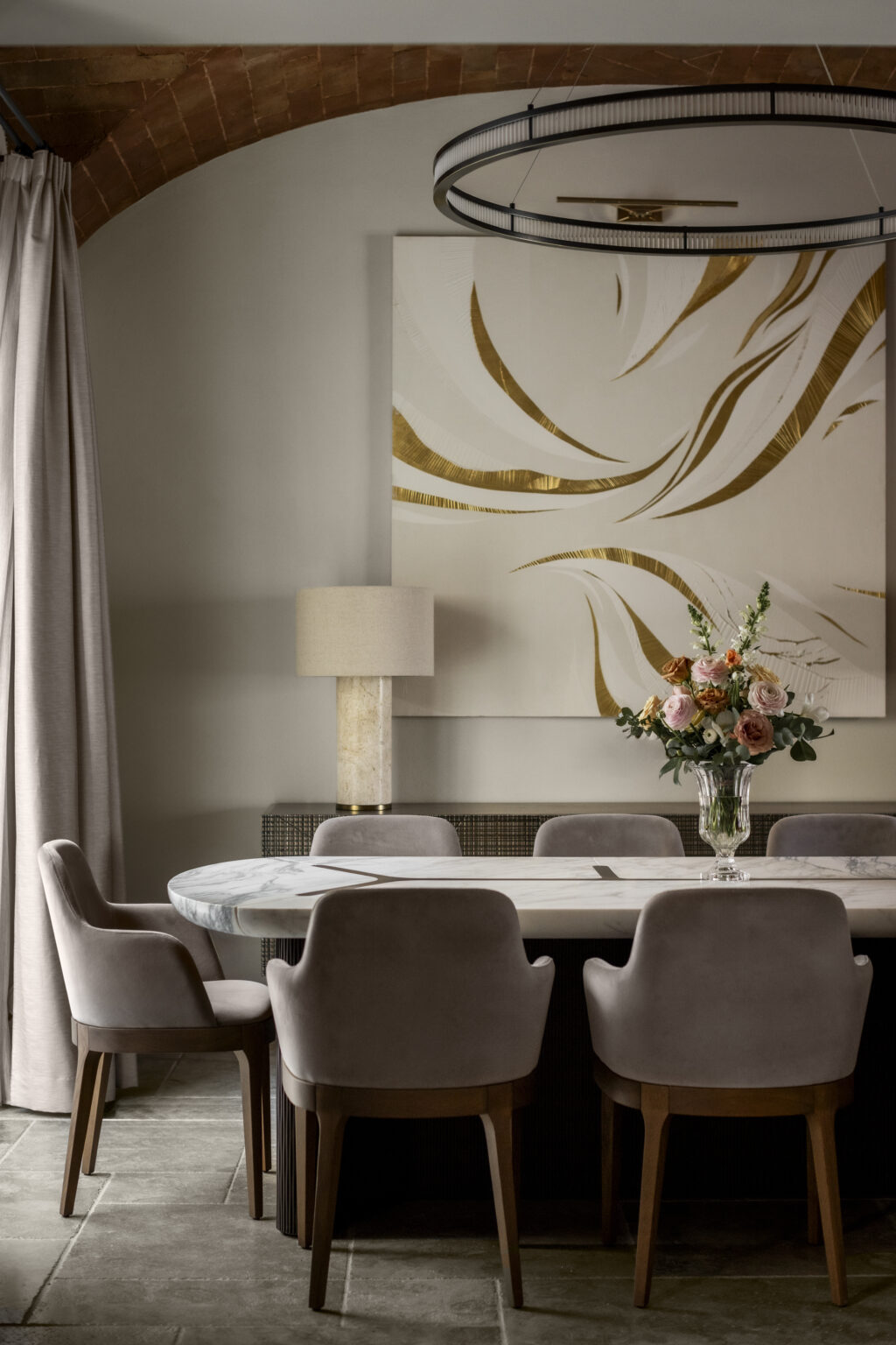 Elegant dining room featuring a marble table surrounded by plush, gray velvet chairs. A modern pendant light hangs above, complementing an abstract gold and white artwork on the wall. A decorative lamp and a vase of colorful flowers add warmth to the sophisticated space. | Only on SopranoVillas