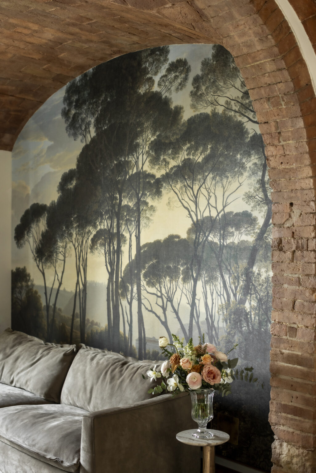 Mural depicting tall trees against a muted sky, showcasing a serene natural landscape, with textured brick wall framing the artwork. | Only on SopranoVillas