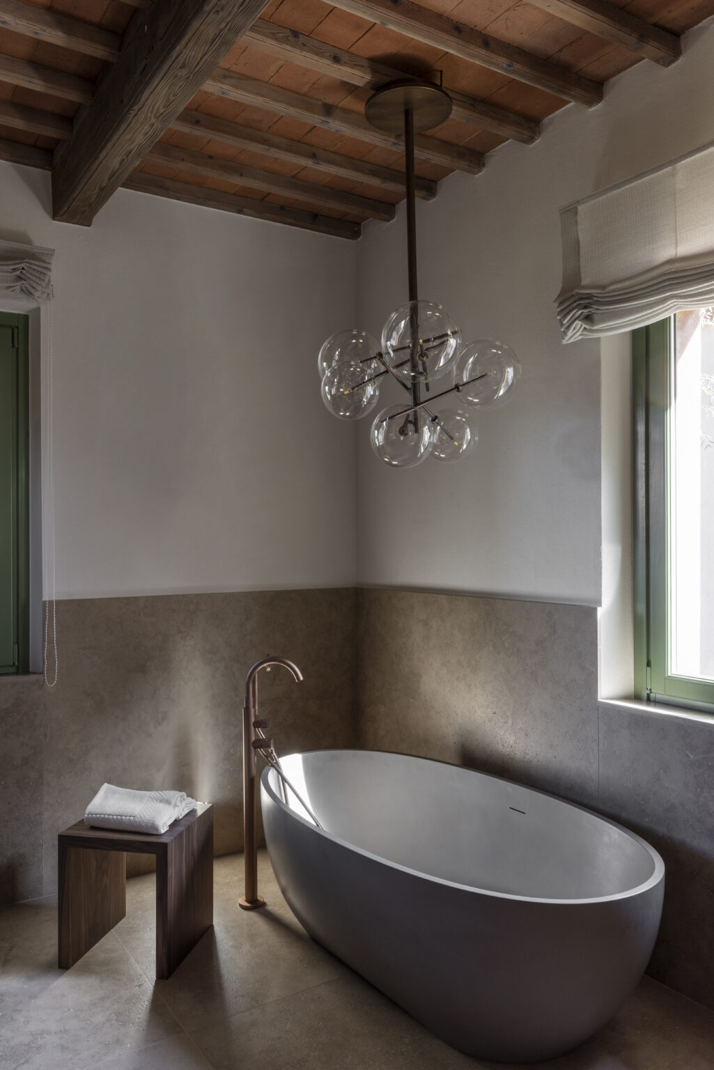 Modern bathroom interior featuring a stylish freestanding bathtub, elegant rose gold faucet, and a contemporary bubble chandelier. Natural light filters through green-framed windows, enhancing the sophisticated design with neutral stone walls. Perfect for inspiration in home decor. | Only on SopranoVillas