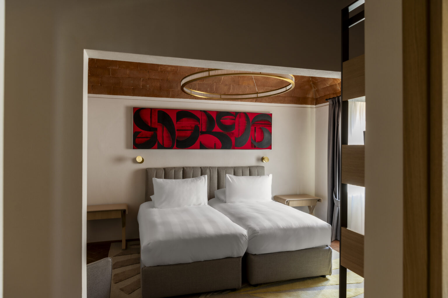 Modern hotel room featuring two twin beds, contemporary decor, and a striking red abstract artwork on the wall, illuminated by a circular light fixture. | Only on SopranoVillas