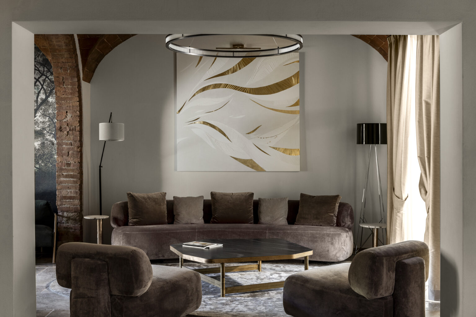 Modern living room featuring a curved brown sofa, two matching armchairs, a glass coffee table, and elegant decor. The room is illuminated by a stylish circular chandelier and features a large abstract painting with gold accents on the wall. Natural light filters through the curtains, highlighting the sophisticated design elements. | Only on SopranoVillas