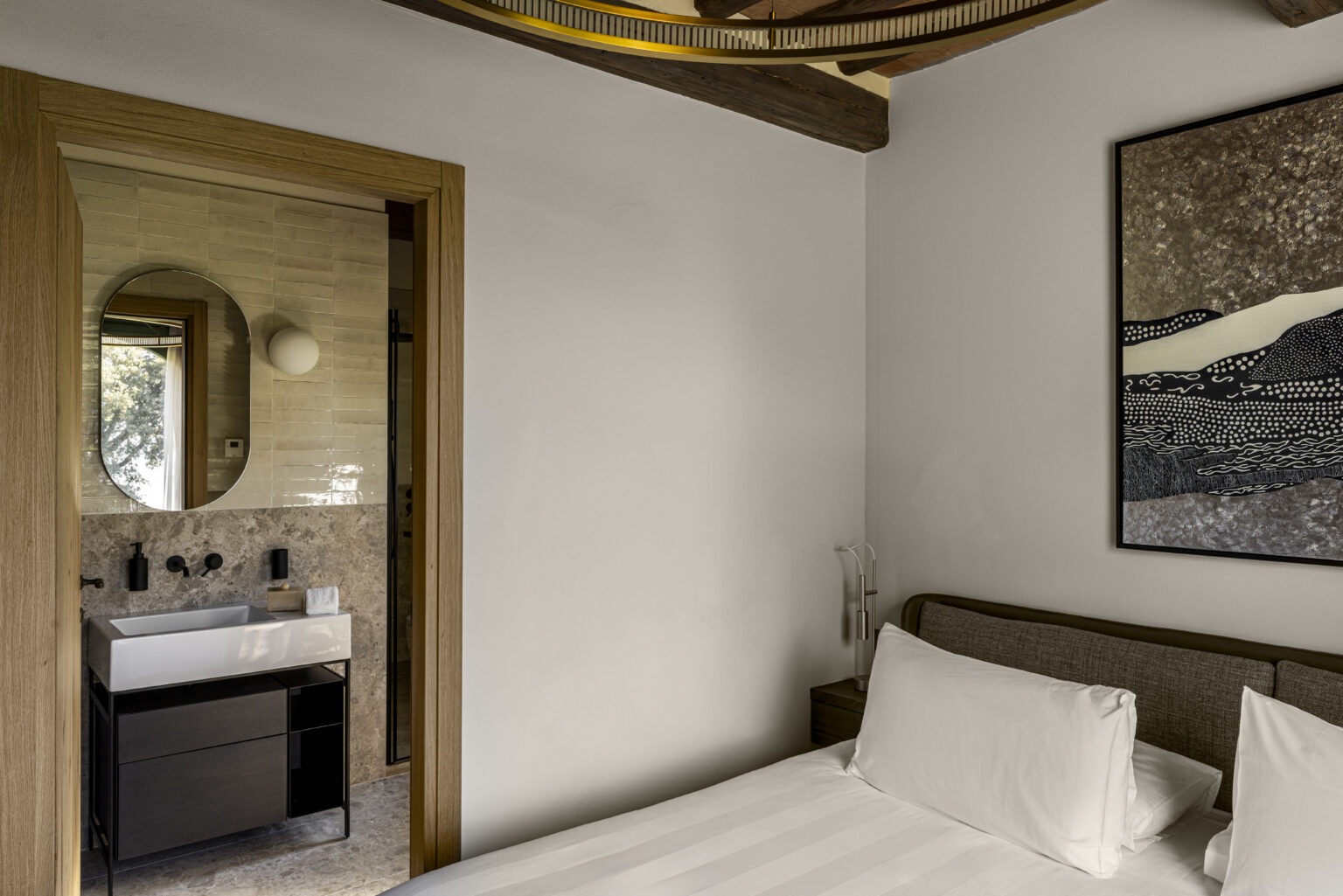 Modern bedroom with a cozy bed, featuring a minimalist design and natural light. The room includes an open doorway leading to a stylish bathroom with a round mirror and sleek vanity. A piece of abstract art hangs on the wall, enhancing the contemporary aesthetic. | Only on SopranoVillas