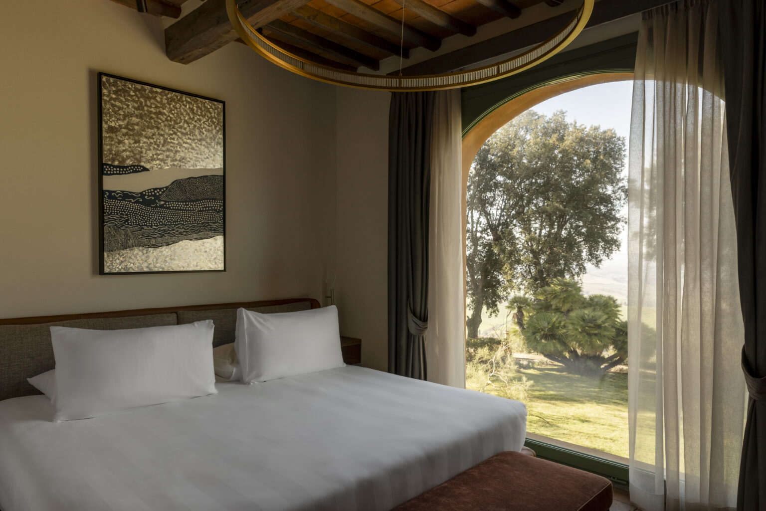 Cozy hotel room featuring a king-sized bed with white linens, a large window offering a view of lush greenery, and modern decor elements including a framed artwork and a stylish light fixture. | Only on SopranoVillas