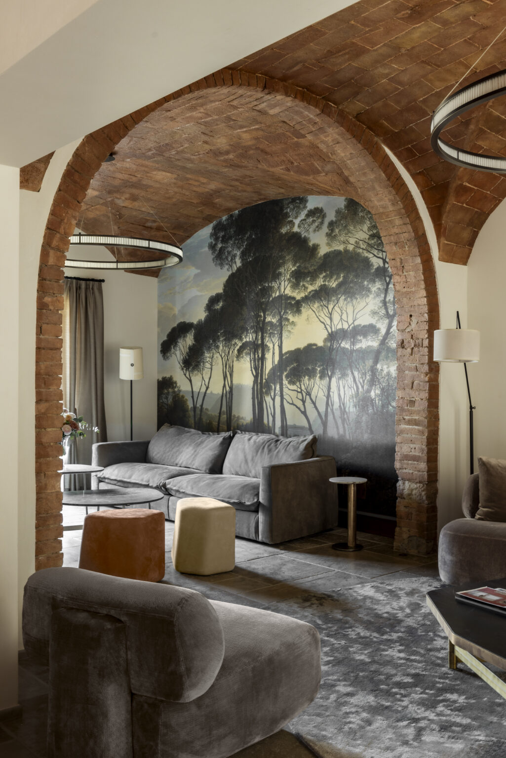 Stylish living room featuring a textured brick archway, a mural of trees on the wall, and modern furniture including a gray sofa, round glass coffee table, and colorful stools. Soft lighting and elegant decor create a cozy atmosphere. | Only on SopranoVillas