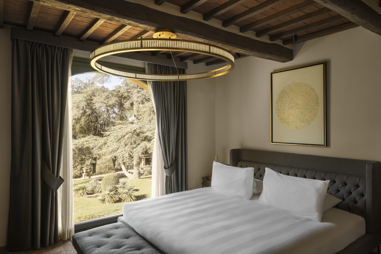 Modern bedroom interior featuring a king-size bed with white linens, elegant gray curtains, and a stylish circular chandelier. A large window offers a view of a landscaped garden, enhancing the serene atmosphere. | Only on SopranoVillas