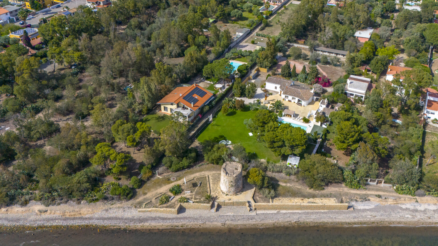 Aerial view of a coastal property featuring a historic tower, surrounded by lush greenery and residential homes, with a sandy beach in the foreground. | Only on SopranoVillas