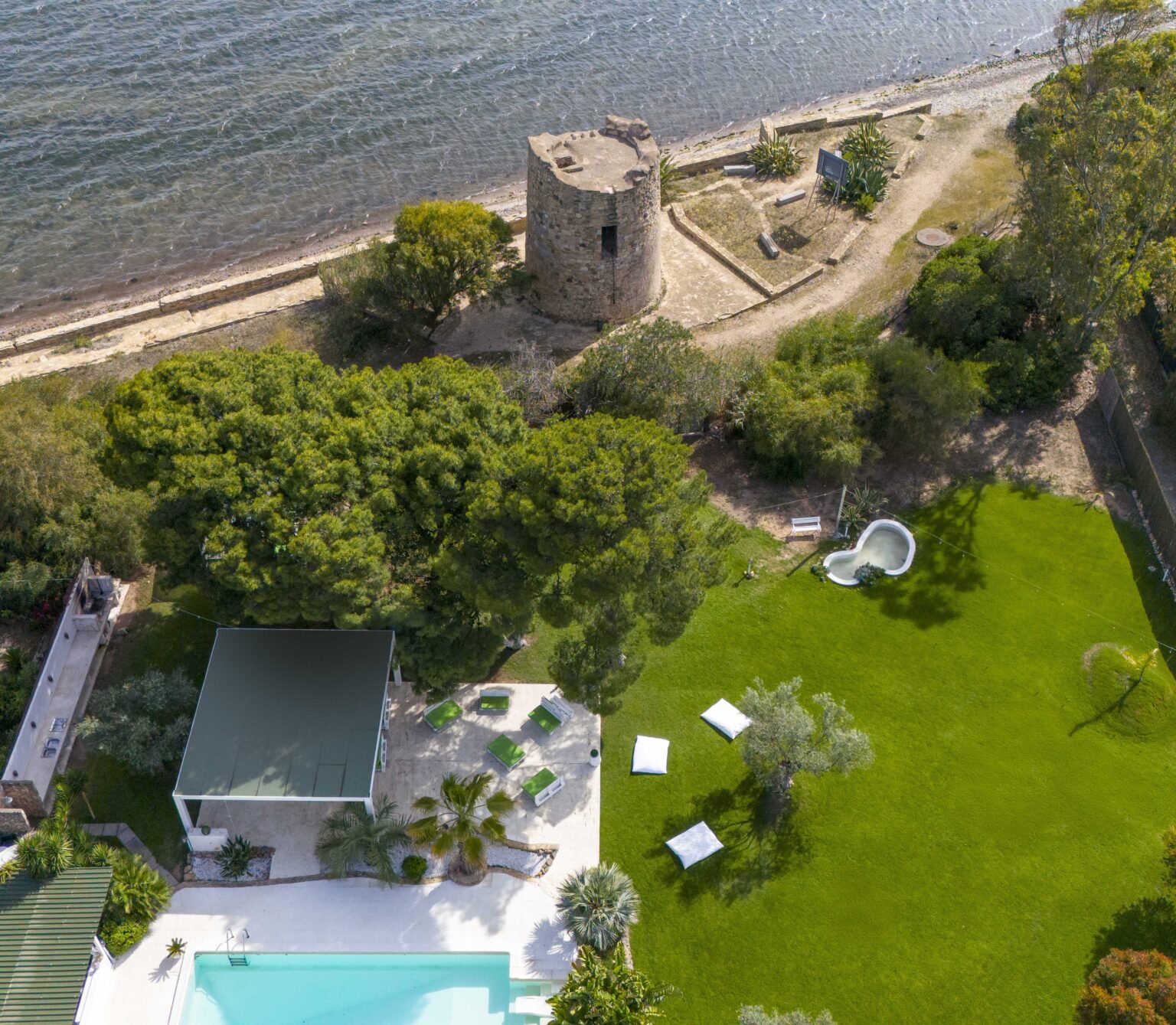 Aerial view of a coastal property featuring a stone tower, lush green lawn, and a swimming pool. The image showcases outdoor lounge areas and scenic waterfront access, highlighting the tranquil landscape and recreational space. | Only on SopranoVillas