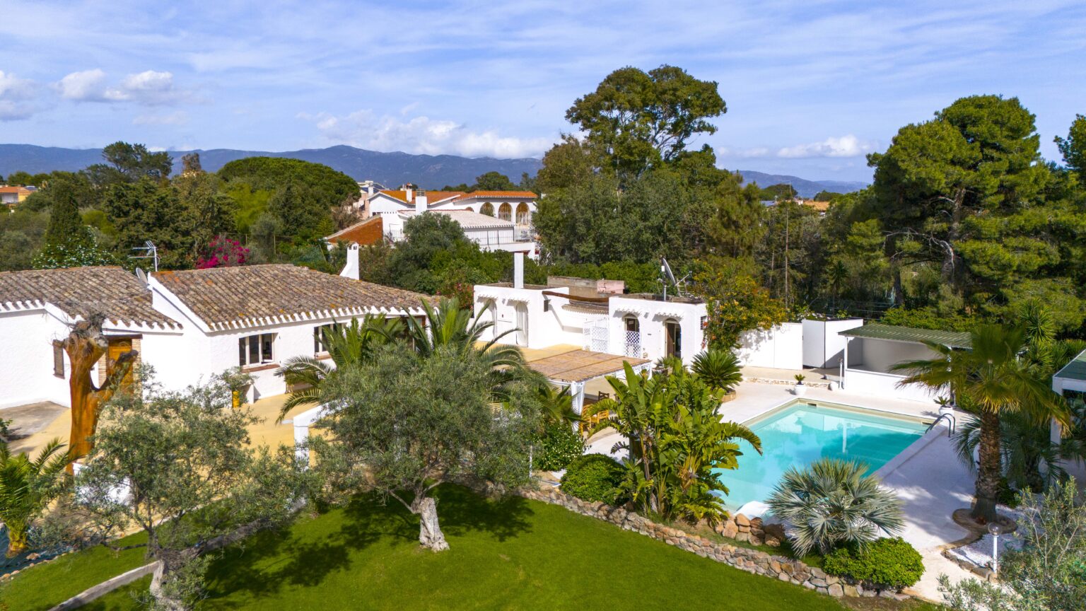 Aerial view of a luxurious villa featuring a private swimming pool, landscaped gardens, and surrounding trees, set against a backdrop of mountains and blue skies. | Only on SopranoVillas