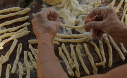 Hands shaping fresh pasta on a wooden surface, surrounded by strips of dough ready for cooking.