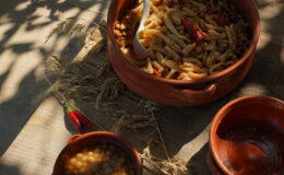Traditional clay pots filled with pasta and spices, with a side of lentil dish, set on a rustic wooden table surrounded by dried herbs.