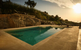 Serene outdoor swimming pool surrounded by natural stone and greenery, with a sunset casting warm light over the scene. Ideal for relaxation and leisure in a tranquil setting.