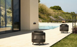 Outdoor patio featuring modern metal fire pits beside a swimming pool, with lounge chairs and lush greenery in the background.
