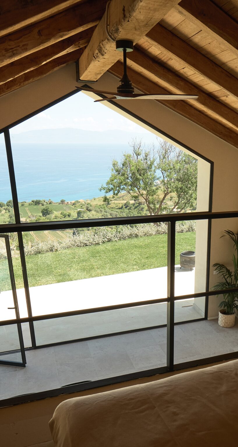 View from a modern interior showcasing a large window overlooking a lush green landscape and the ocean beyond, with a potted plant in the foreground. | Only on SopranoVillas