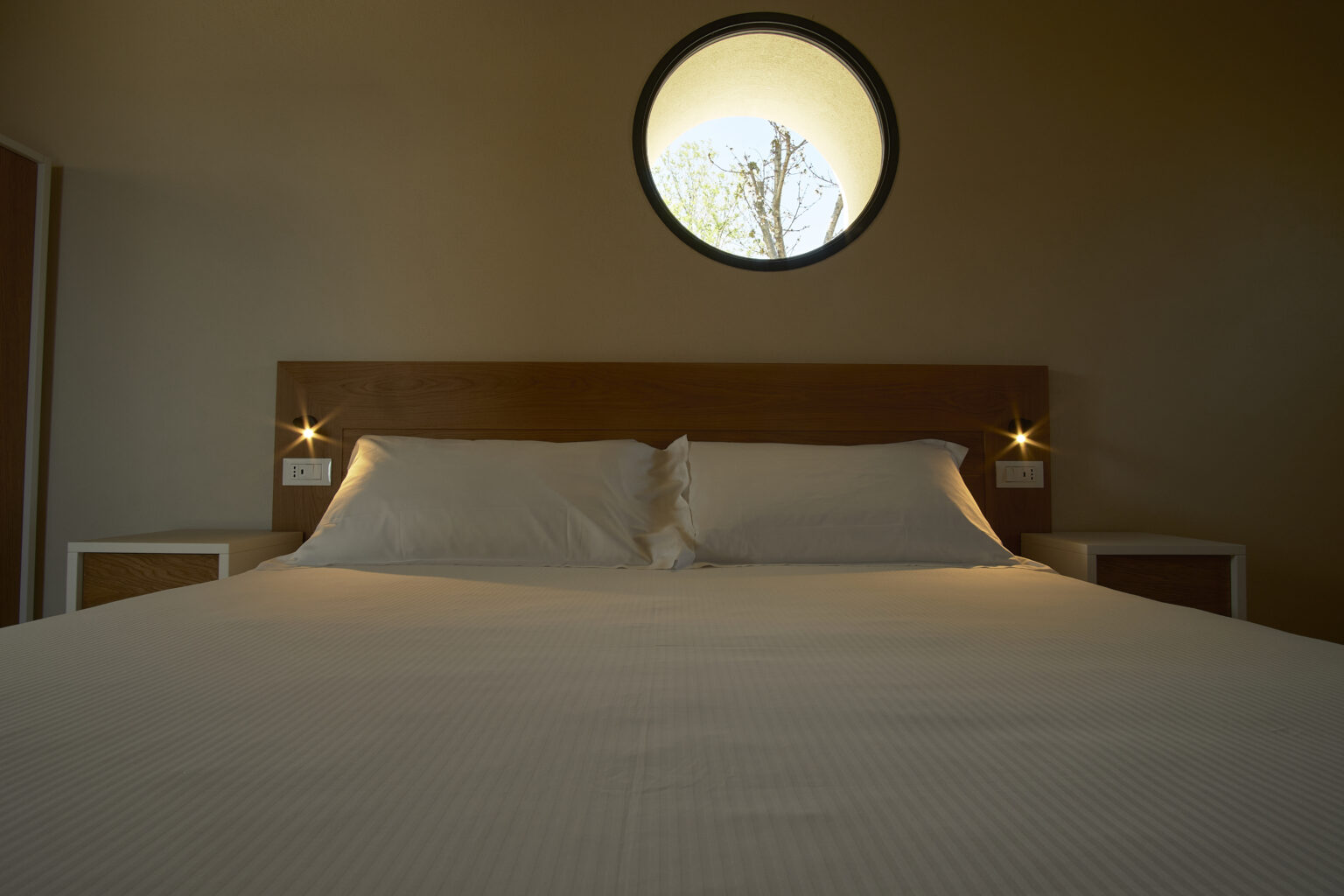 Modern bedroom interior featuring a wooden bed frame, white bedding, and bedside lamps, with a round window offering a view of trees outside. | Only on SopranoVillas
