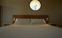 Modern bedroom interior featuring a wooden bed frame, white bedding, and bedside lamps, with a round window offering a view of trees outside.