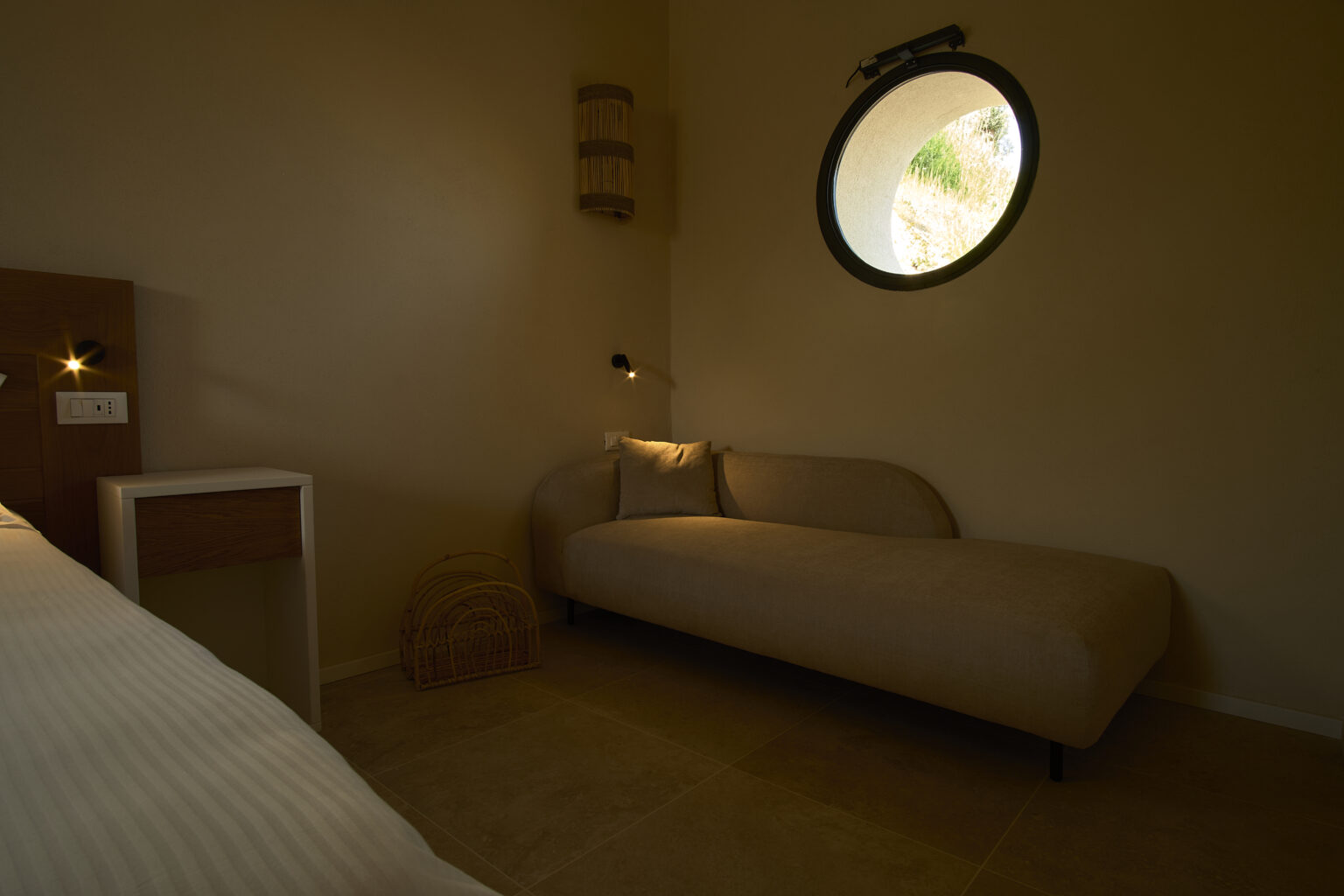 Cozy bedroom interior featuring a minimalist design, a circular window allowing natural light, a comfortable daybed, and a bedside table with modern lighting. | Only on SopranoVillas