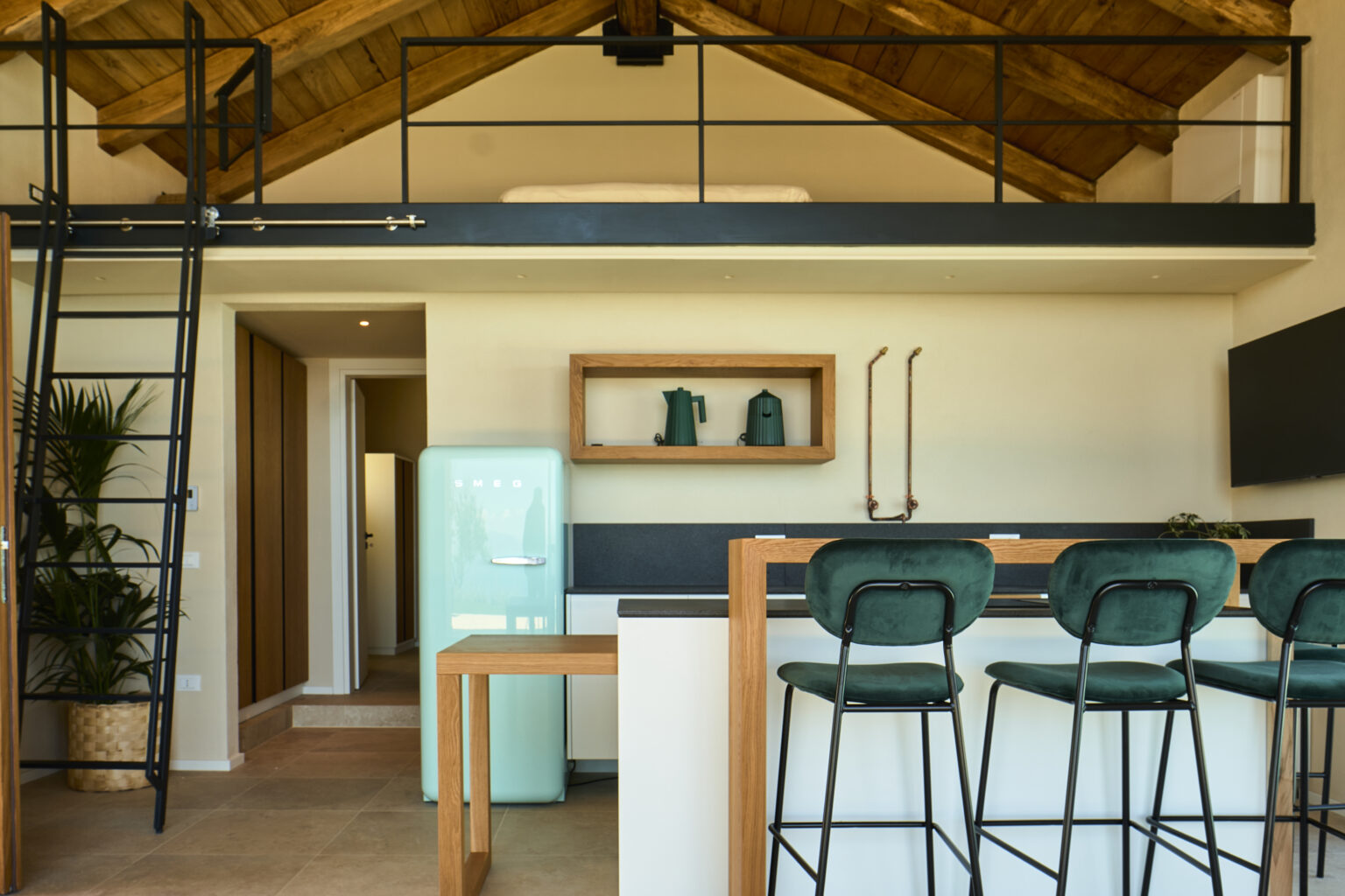 Modern kitchen interior featuring a loft design, with a green retro refrigerator, stylish bar stools, and a sleek countertop. The space is illuminated by natural light, showcasing wooden beams and contemporary decor elements. | Only on SopranoVillas